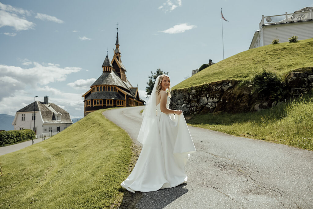 Bride walking toward St. Olaf’s Church in Balestrand, Norway, turning back to look into the camera with the church visible in the background.
